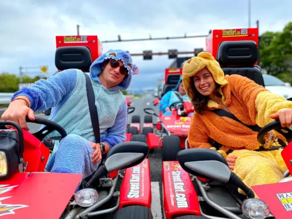 Two people in blue and orange fuzzy animal costumes sit in red go-karts on a road, smiling at the camera.